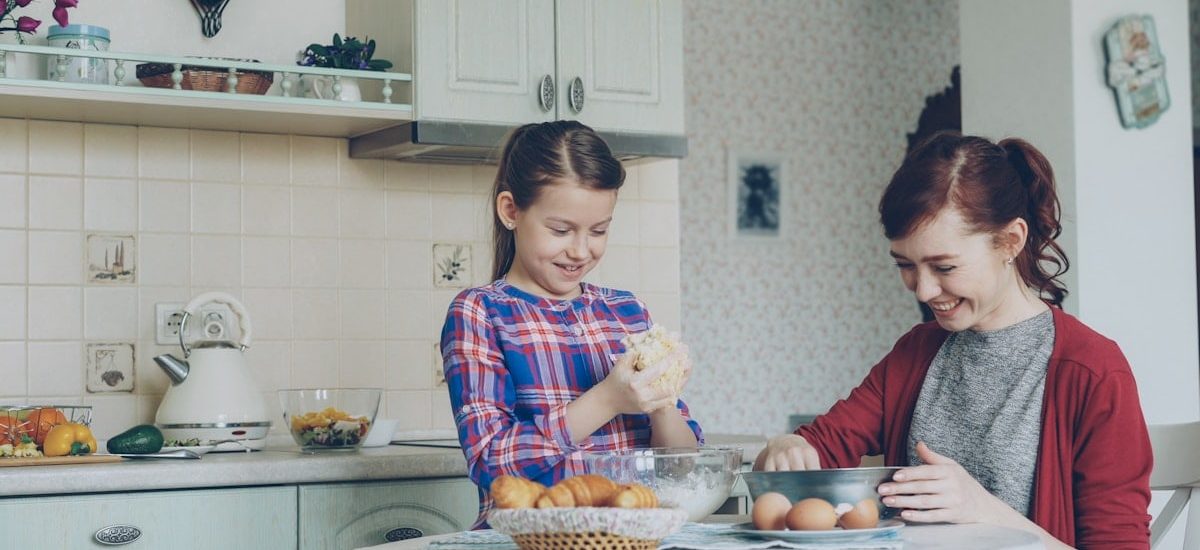 Little cute girl helping her mother in the kitchen stirring dough for cookies into bowl. Mom have fun smearing daughter nose with flour and they laughing . Family, food, home and people concept