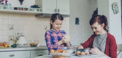 Little cute girl helping her mother in the kitchen stirring dough for cookies into bowl. Mom have fun smearing daughter nose with flour and they laughing . Family, food, home and people concept