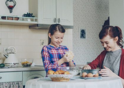 Little cute girl helping her mother in the kitchen stirring dough for cookies into bowl. Mom have fun smearing daughter nose with flour and they laughing . Family, food, home and people concept